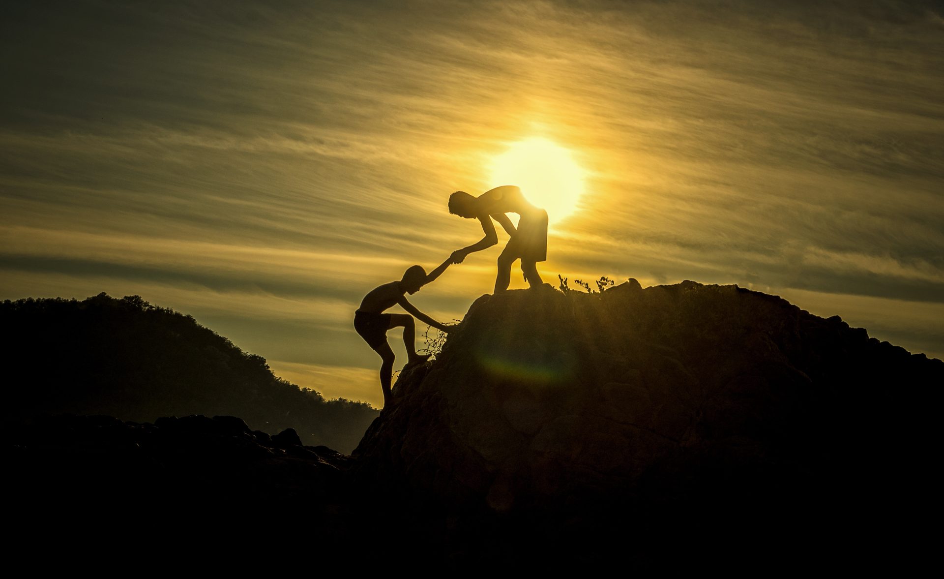 One person reaching down to help another climb a rocky cliff at sunset